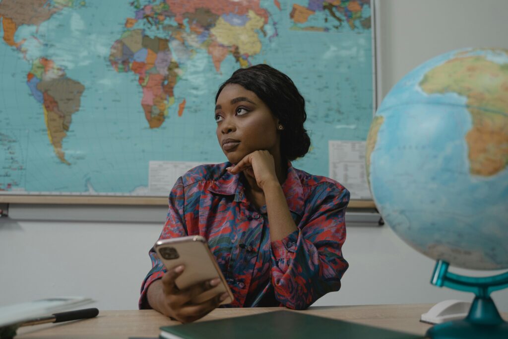 A teacher in a classroom, pondering with a smartphone. Educational setting with a globe and map.