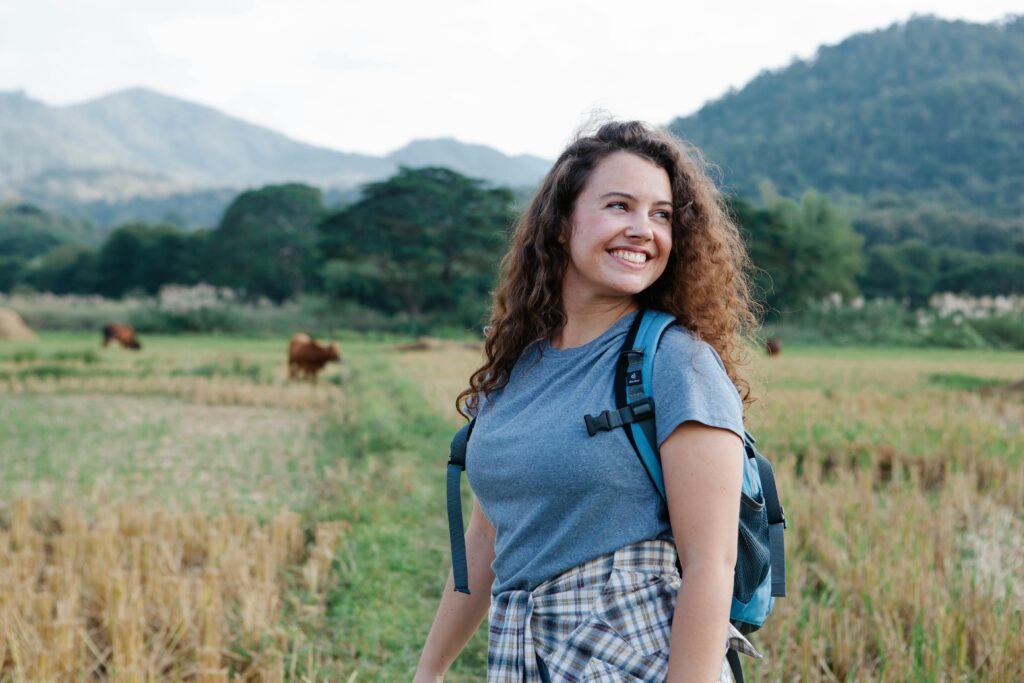 Woman with backpack enjoys a peaceful hike through lush countryside with mountains and grazing cows.