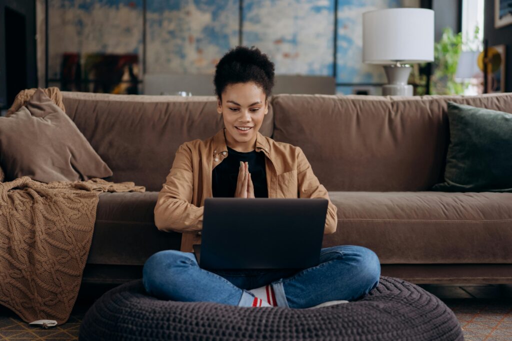 A smiling young woman sits cross-legged on a pouf, using her laptop in a cozy home setting.