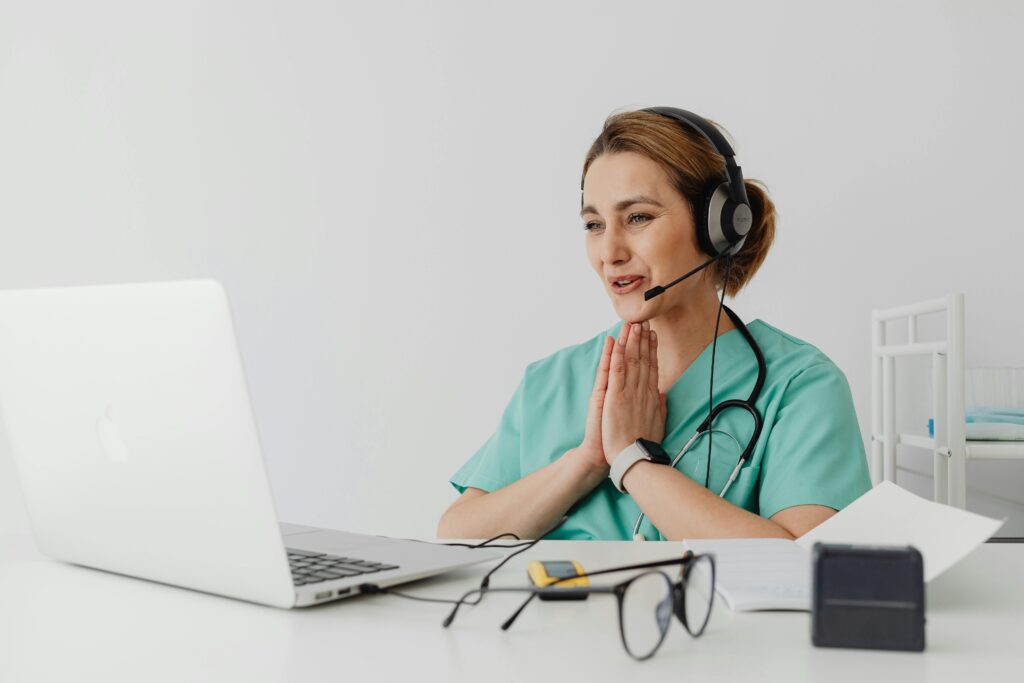 Female nurse using telemedicine for a virtual consultation on a laptop, wearing headphones and stethoscope.