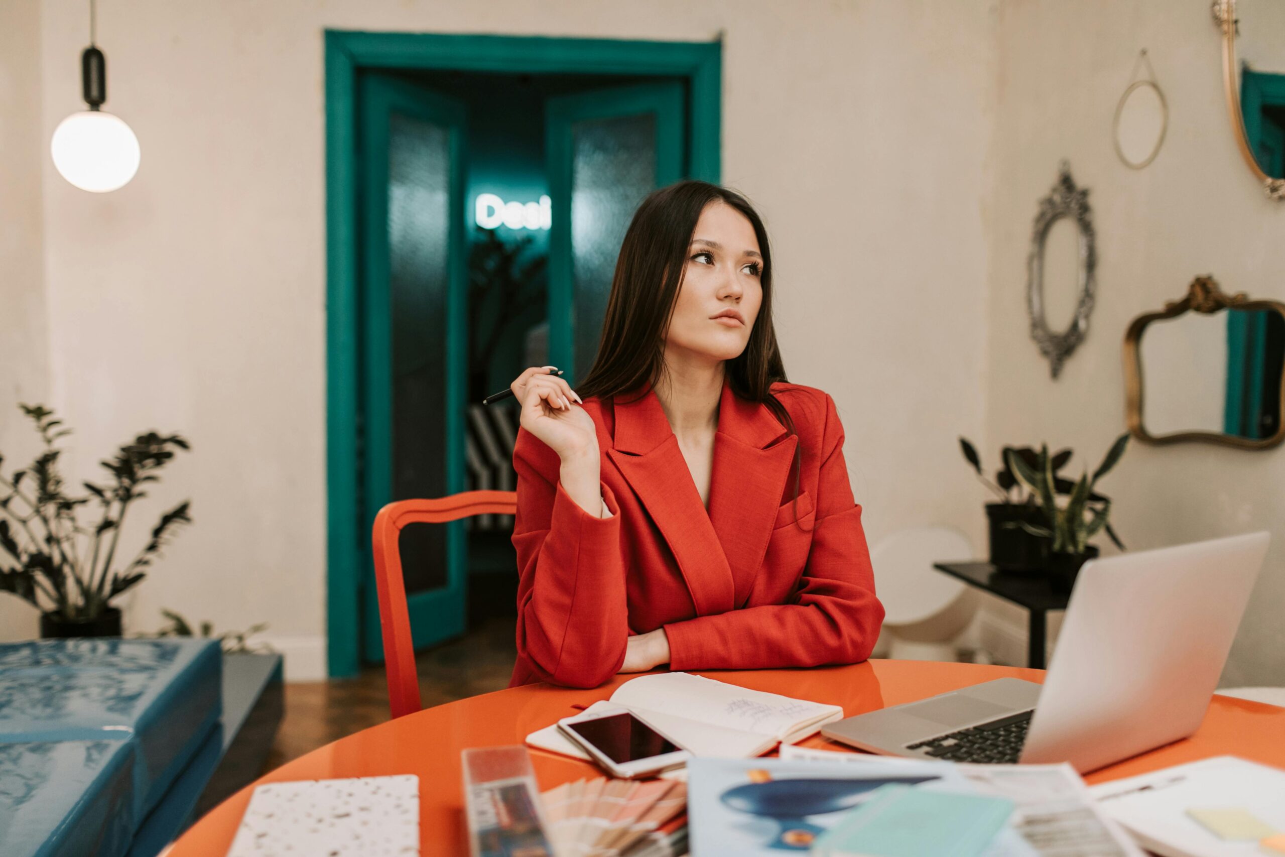 A businesswoman in a red suit sits thoughtfully at an office desk with a laptop, highlighting modern work life.