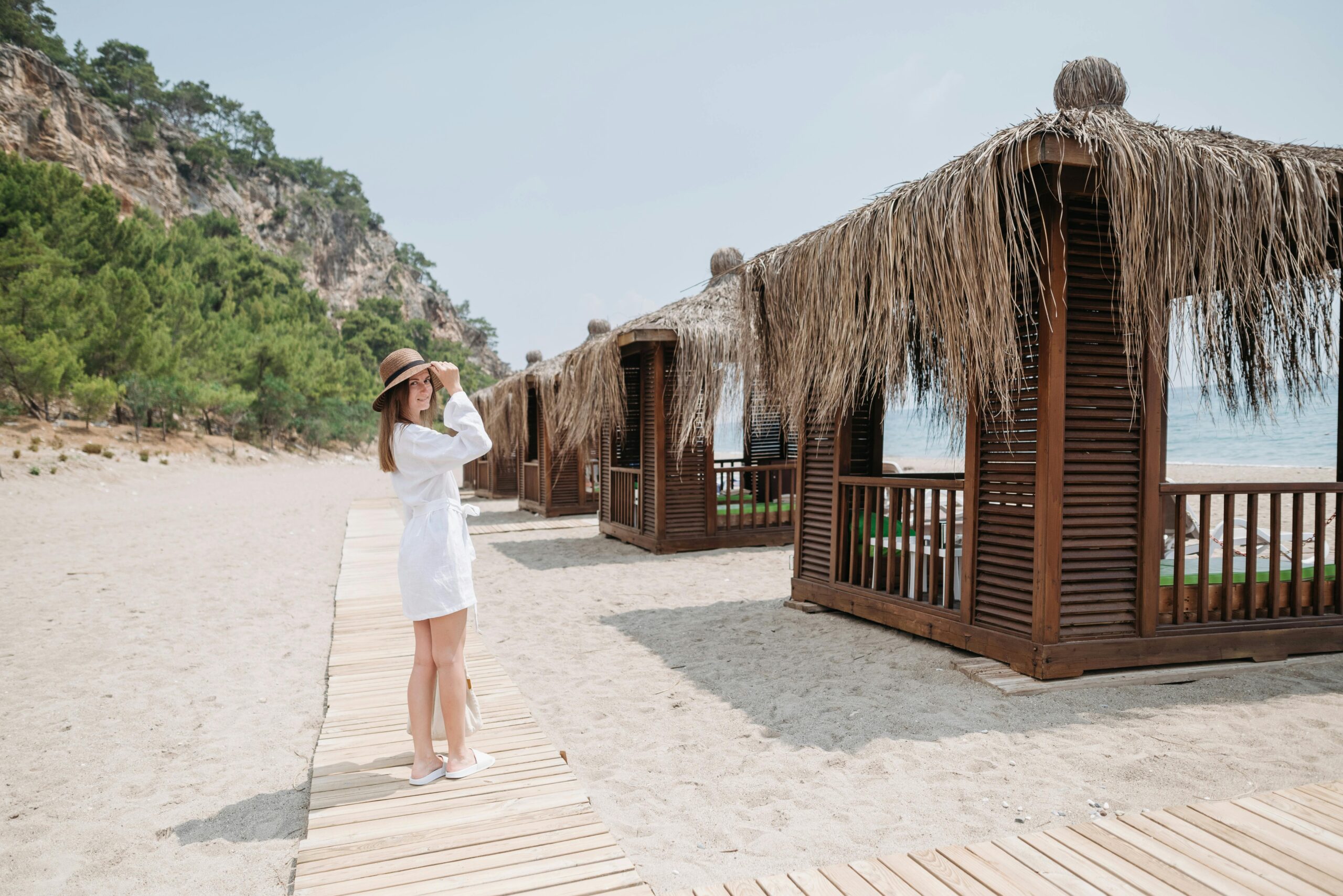 Woman in white dress and hat stands on sandy beach, gazing at scenic wooden cabanas under clear summer sky.