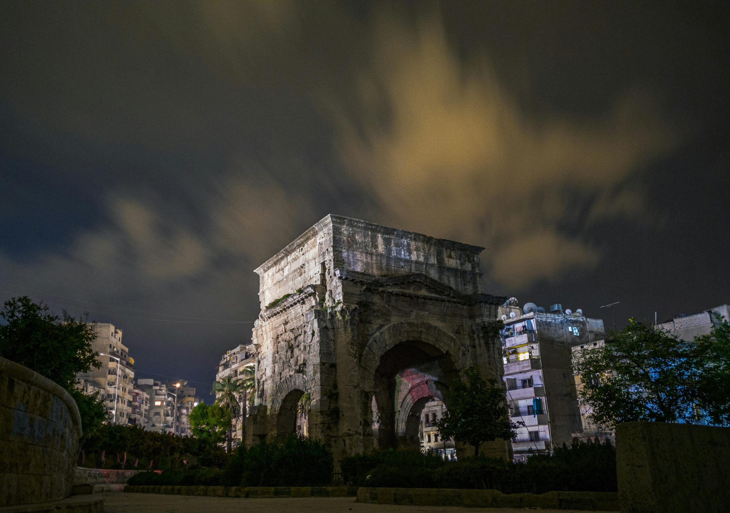 A historic arch in Latakia, Syria illuminated at night with cloudy skies.