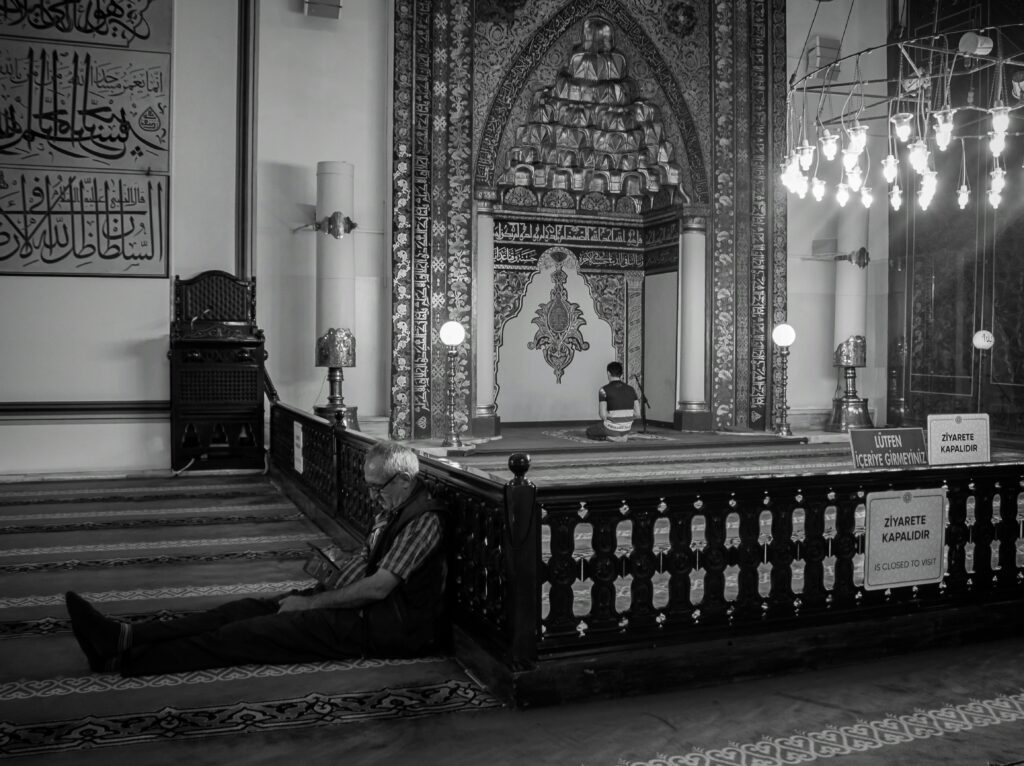 Black and white capture of a mosque interior with individuals engaged in prayer and reflection.