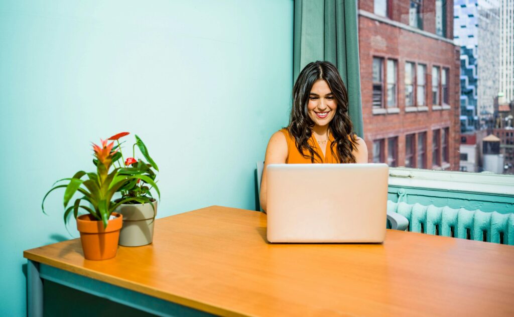 Confident woman in a bright office working on a laptop, surrounded by urban scenery and greenery.