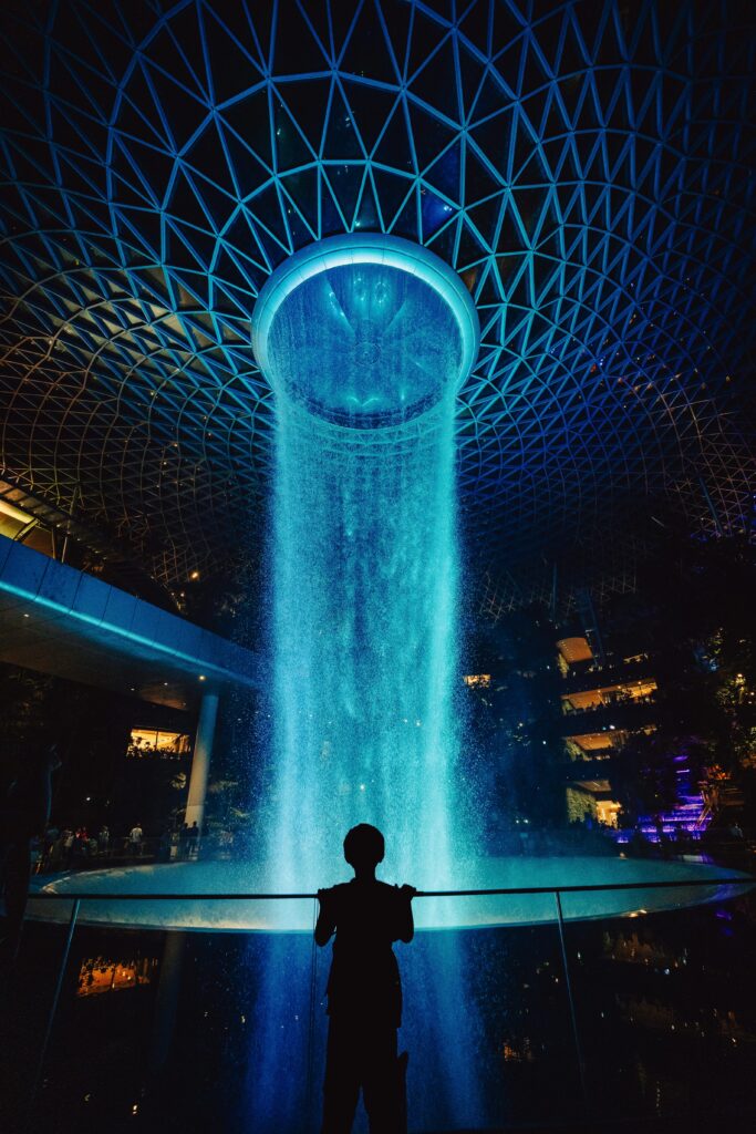 Silhouette of a person admiring the illuminated Vortex Waterfall at Jewel Changi Airport in Singapore.