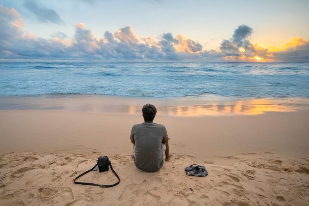 Man sitting on sandy beach at sunset, gazing at the ocean, reflecting a peaceful sunset.