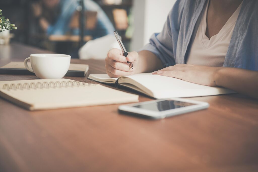 A woman writes in a notebook at a café table with a coffee and smartphone nearby.