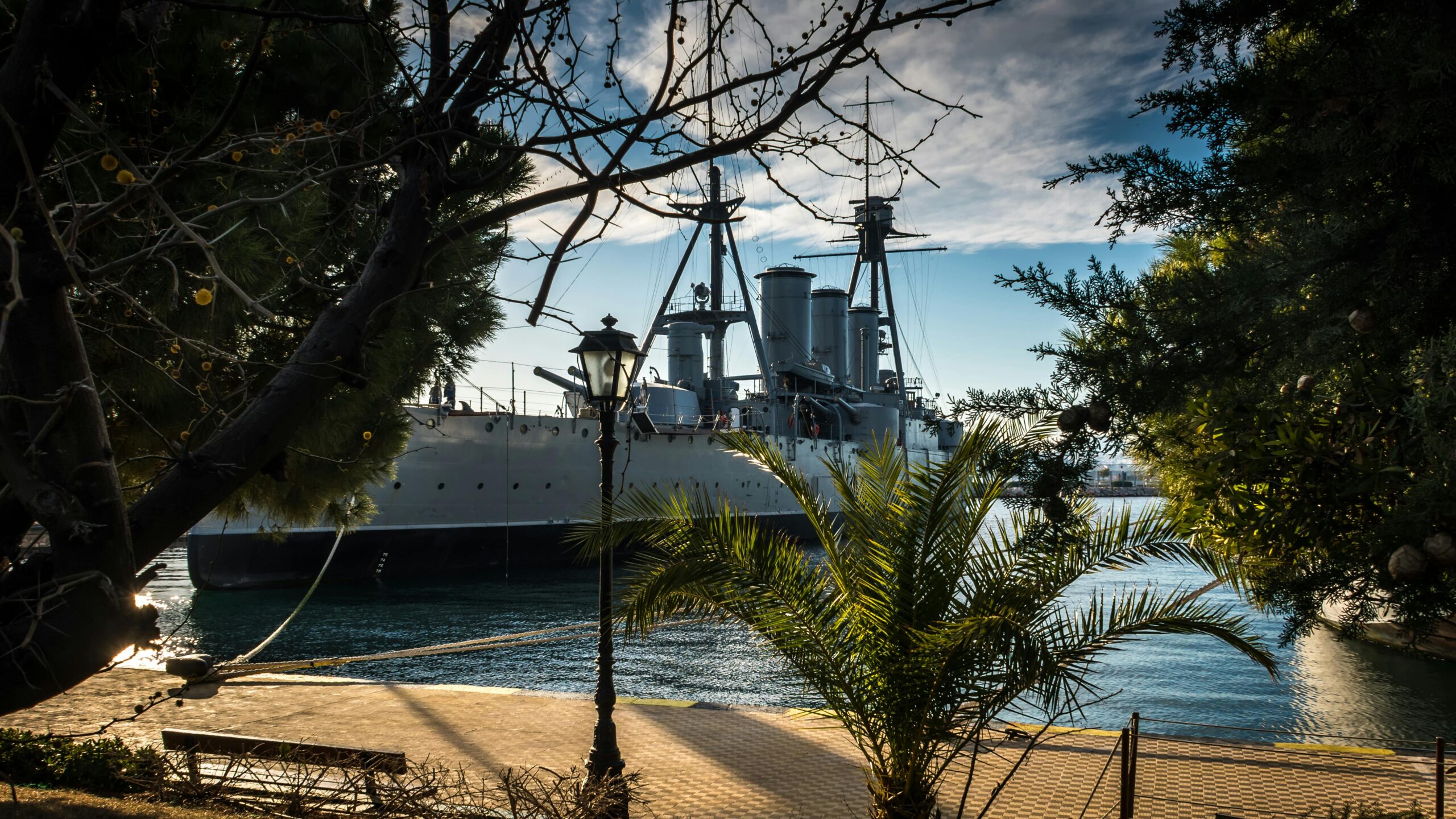 Historic battleship docked at a serene marina, framed by palm trees and a classic lamppost.