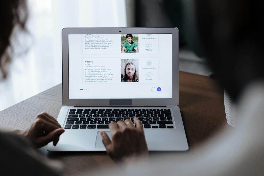 Hands using laptop displaying online profiles of children for adoption, indoors.