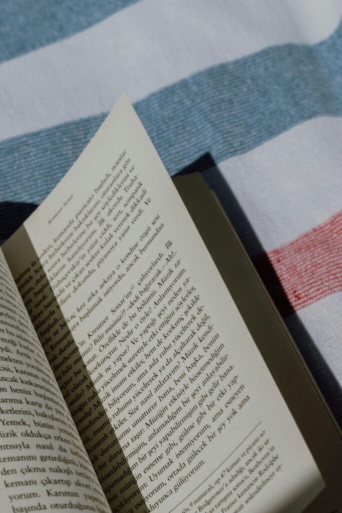 An open book with text in Turkish lies on a striped fabric in natural sunlight.
