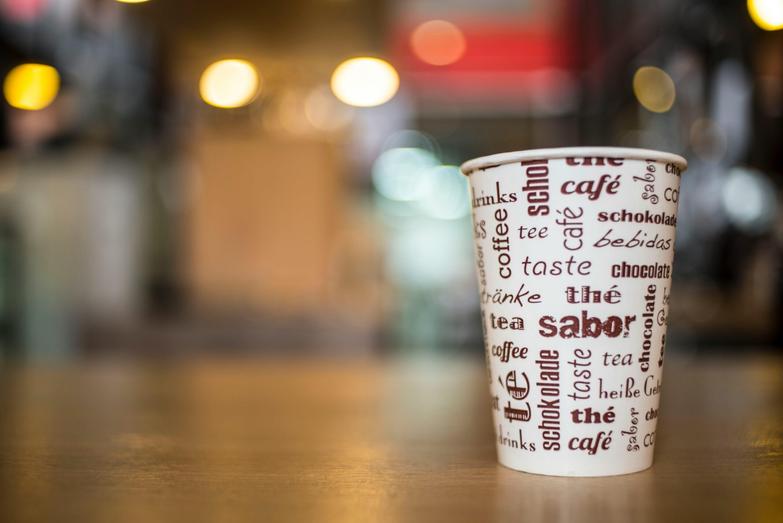 A coffee cup with multilingual text stands on a wooden table in a vibrant café setting.