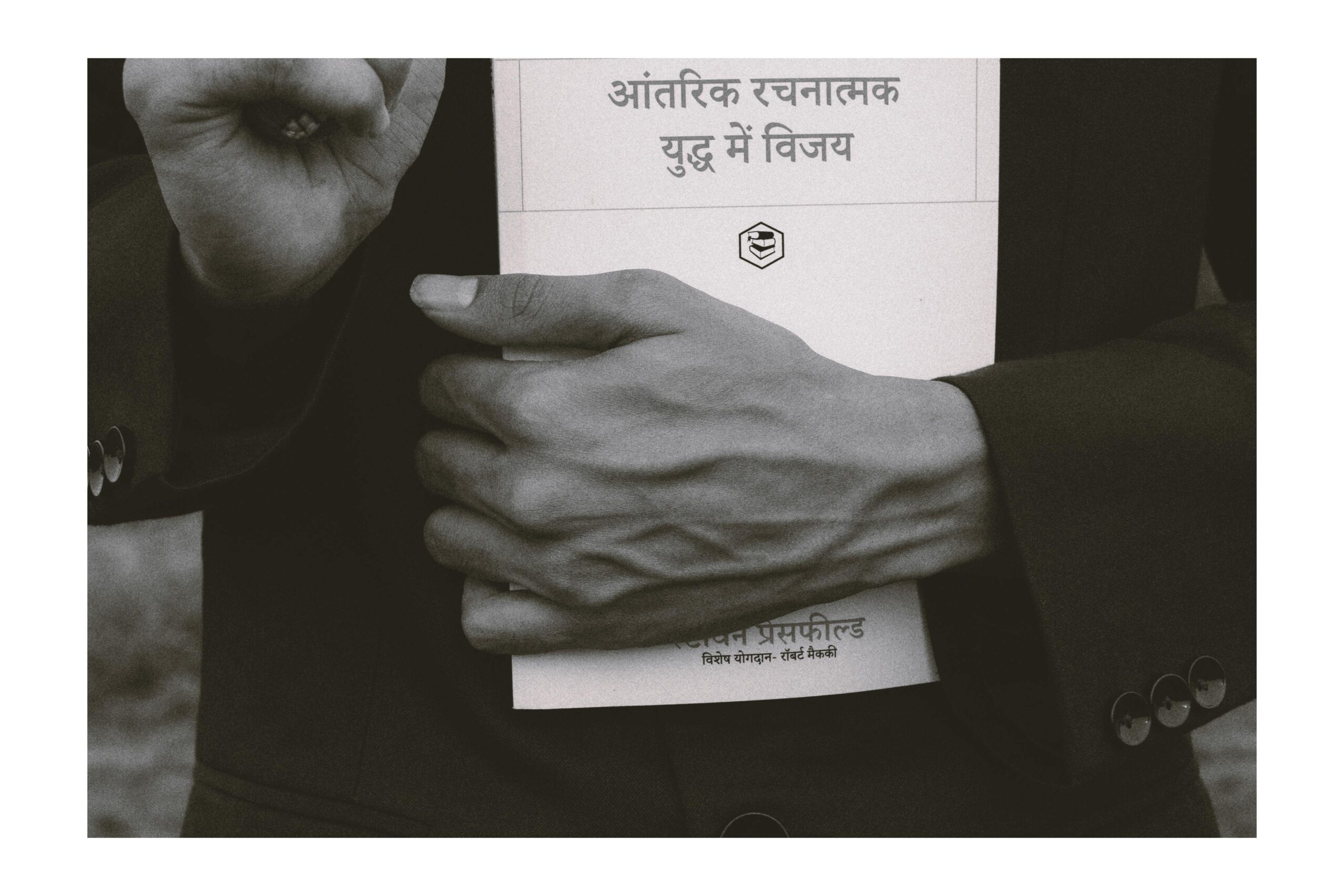 Close-up black and white image of hands holding a book with Hindi text, conveying a thoughtful mood.
