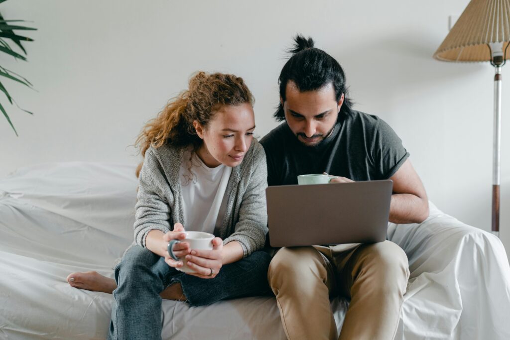 Positive young couple in casual clothes sitting on sofa with white cover and surfing netbook