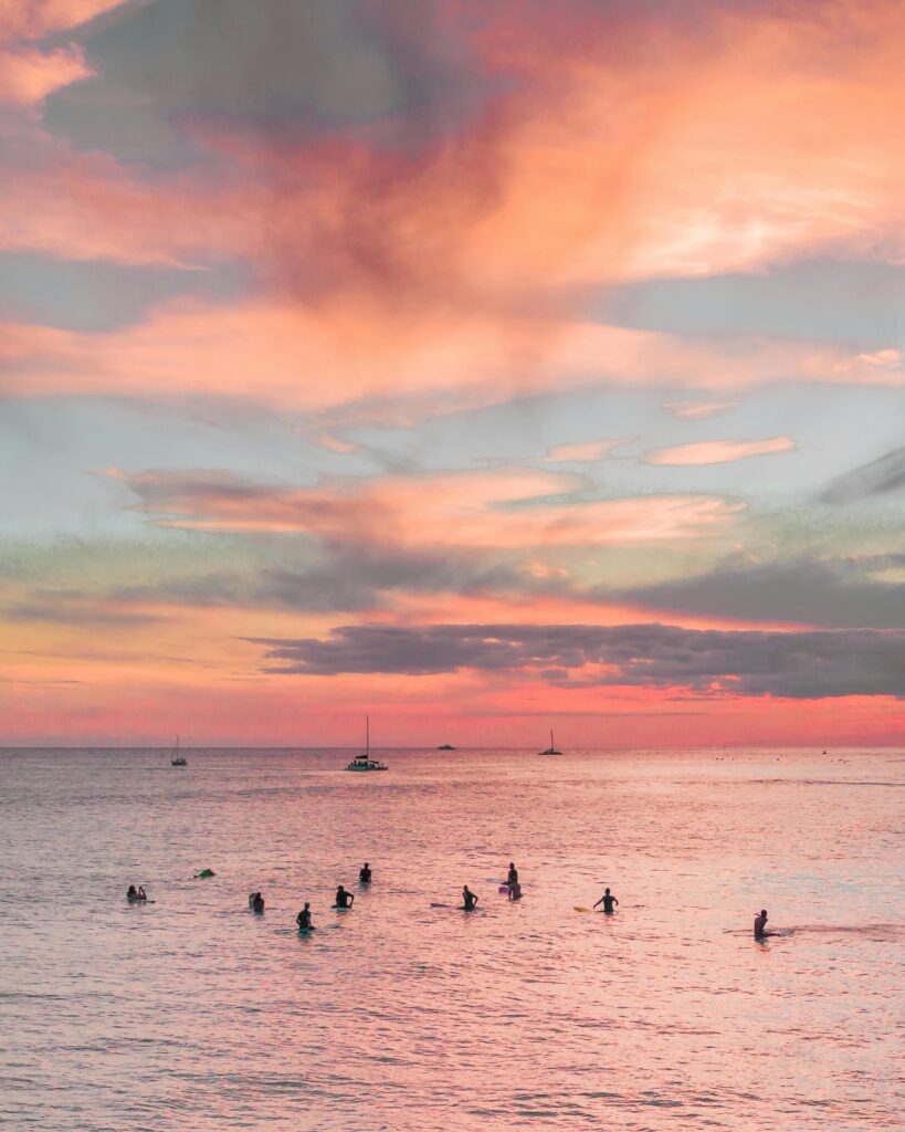 Surfers enjoy a vibrant sunset near Waikiki, reflecting hues of pink and orange over the ocean.