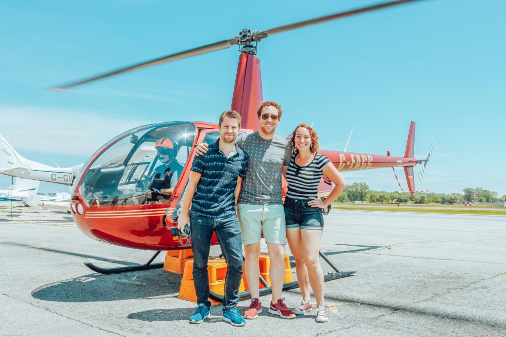Three friends smiling in front of a red helicopter at the airfield, enjoying a sunny day.