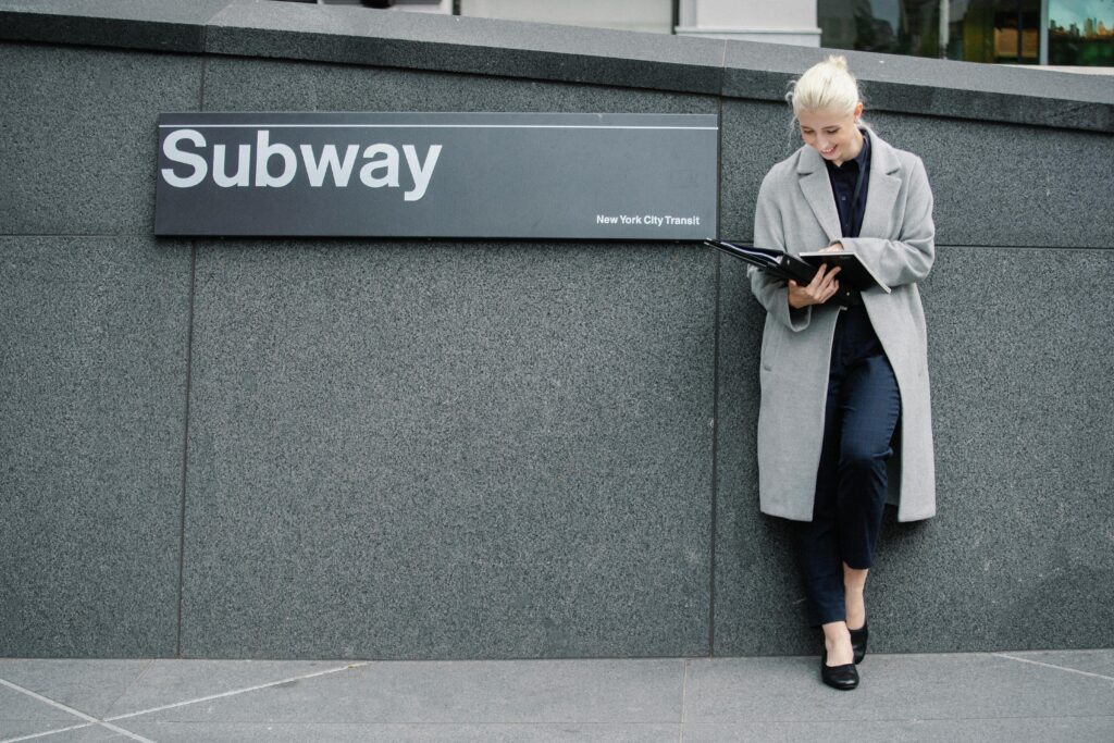 Full body smiling young businesswoman in formal clothes and coat reading documents and standing near subway station entrance