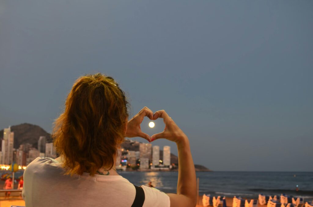 A person forms a heart shape with hands framing the moon over Benidorm beach at night.
