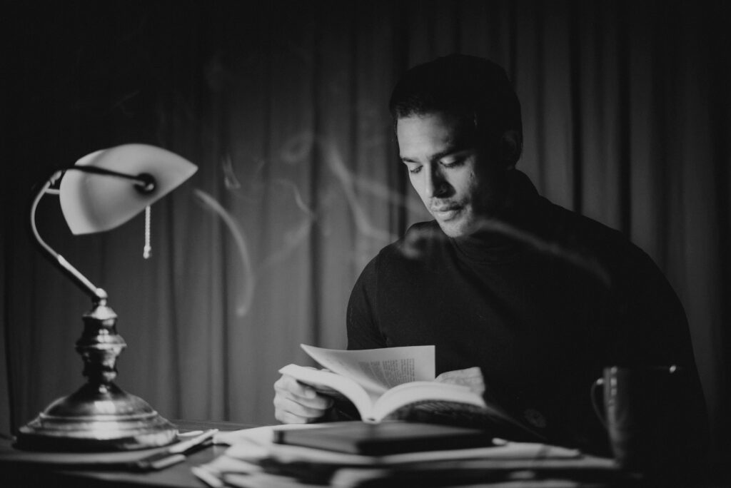 Black and white focused good looking male in casual wear reading book while sitting at desk with papers and bankers lamp in home office