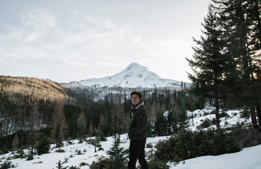 A young man stands amidst a snowy landscape with Oregon's iconic Mount Hood in the background, capturing the essence of winter adventure.