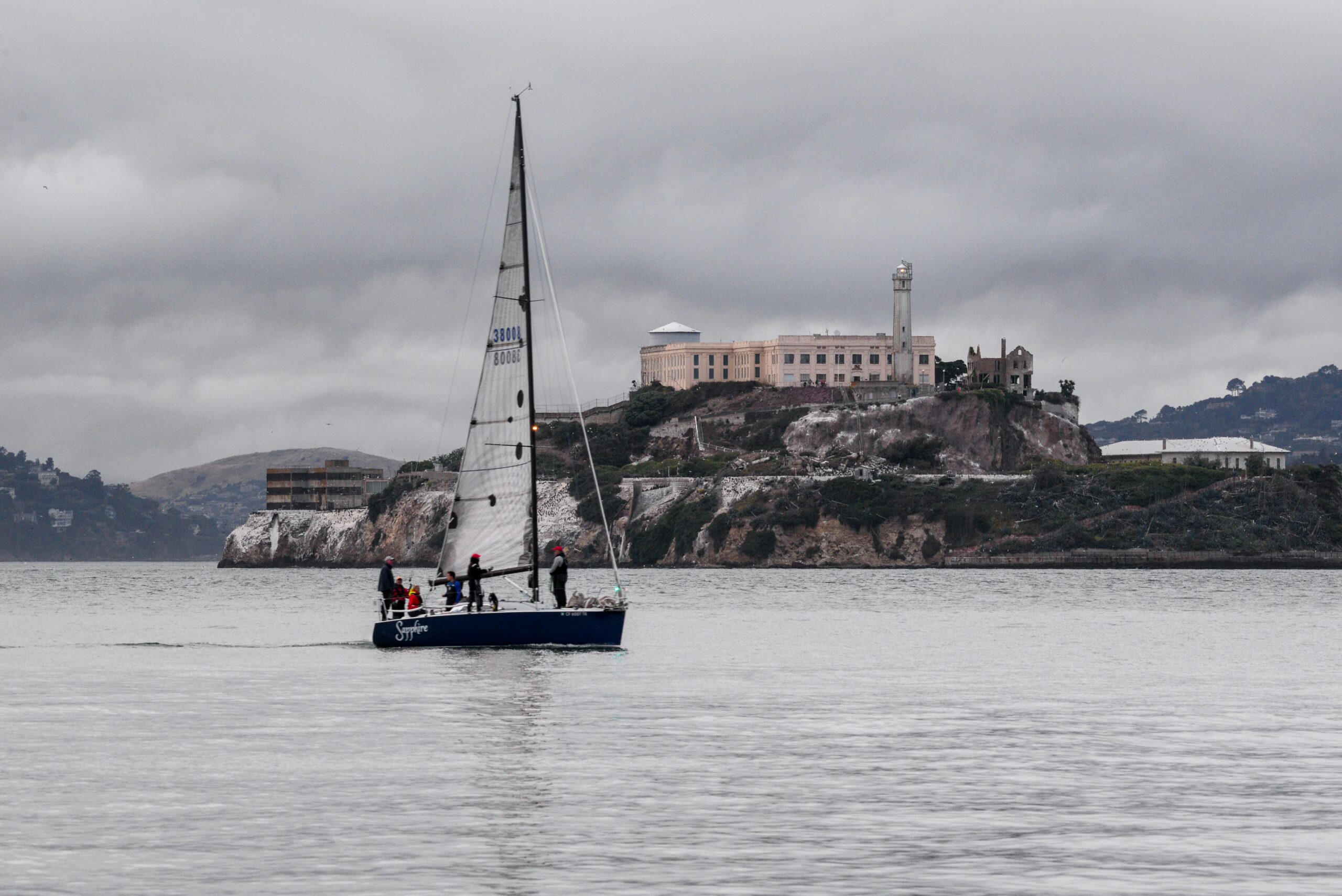 A sailboat cruises near the iconic Alcatraz Island under a cloudy sky in San Francisco Bay.