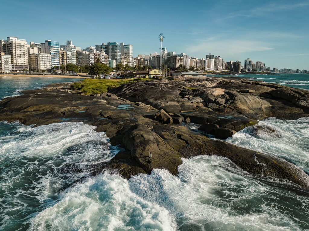 A scenic view of Guarapari's beachfront with crashing waves and city skyline in the background.