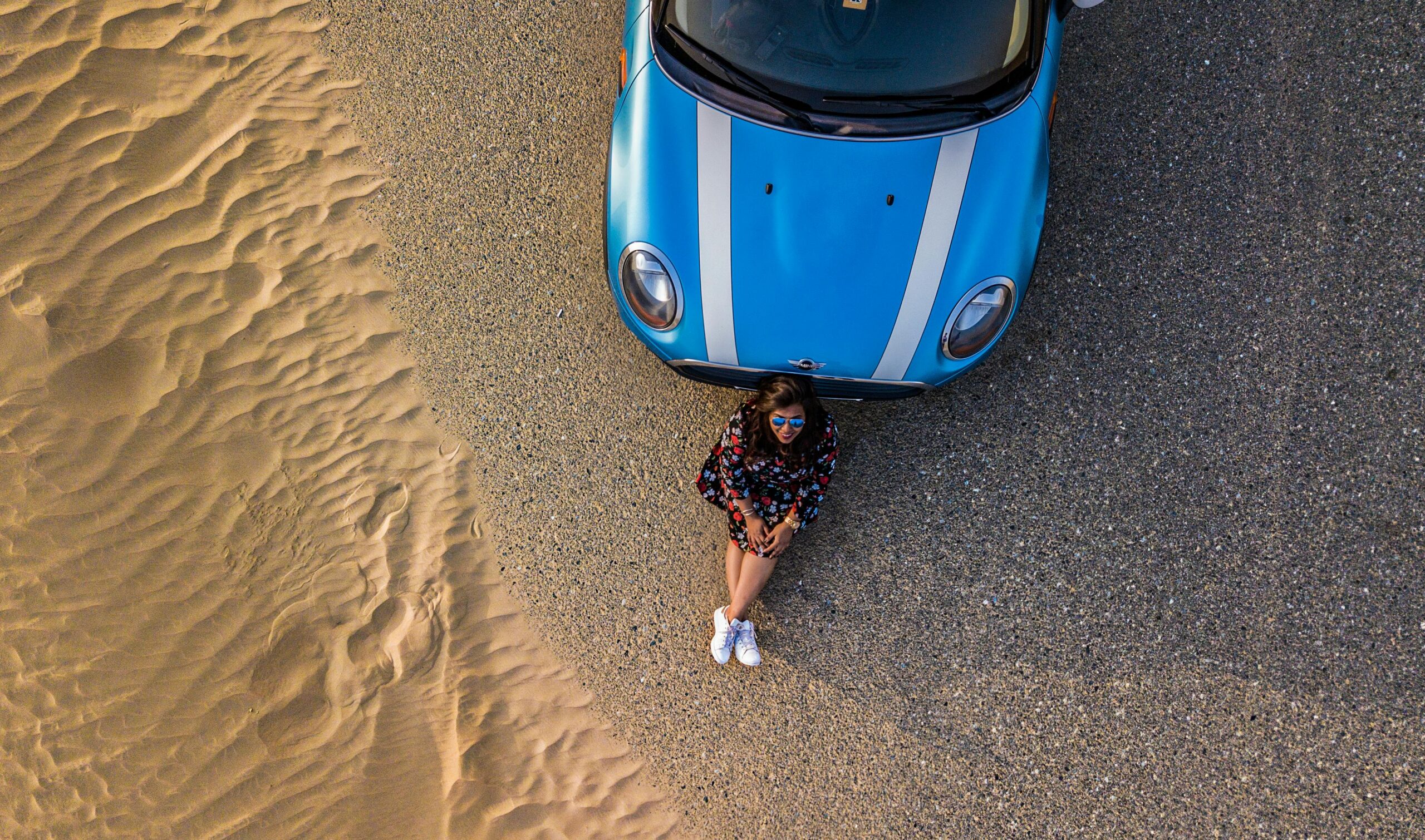 Drone shot of a woman sitting by a blue car on desert sands, enjoying solitude and adventure.