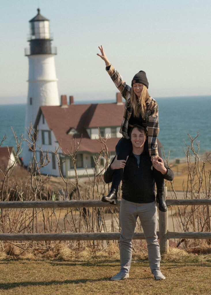 Playful couple pose at Portland Head Light, Portland ME, capturing a carefree moment by the sea.