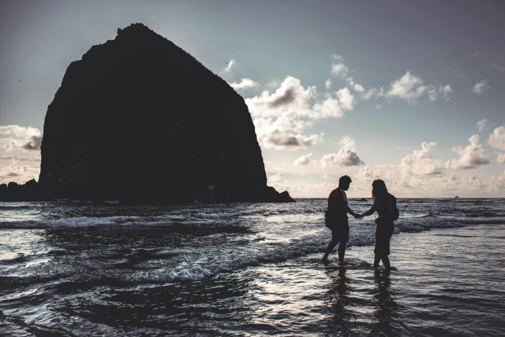 Silhouette of a couple holding hands at Cannon Beach during sunset, with Haystack Rock in view.