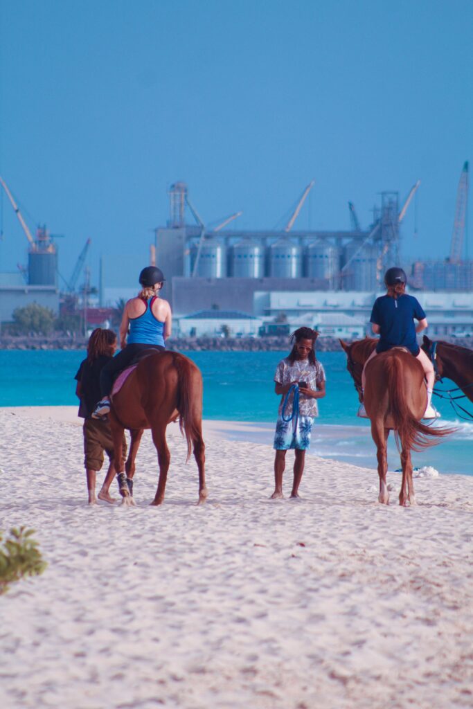 People horseback riding on a sandy beach with an industrial skyline in the background.