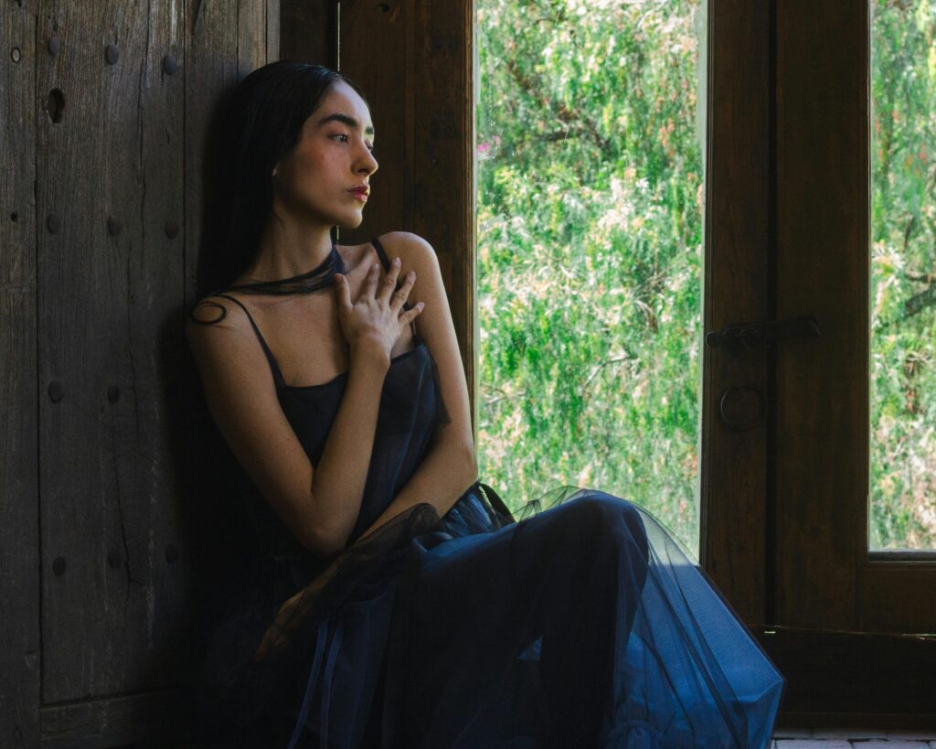 A woman in a dark dress sits thoughtfully by a window, capturing a serene and introspective moment.