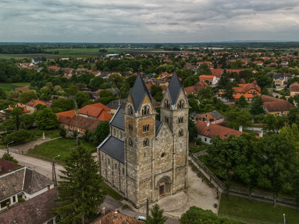 Aerial view of a Romanesque church surrounded by lush greenery and red-roofed houses in a Hungarian village.