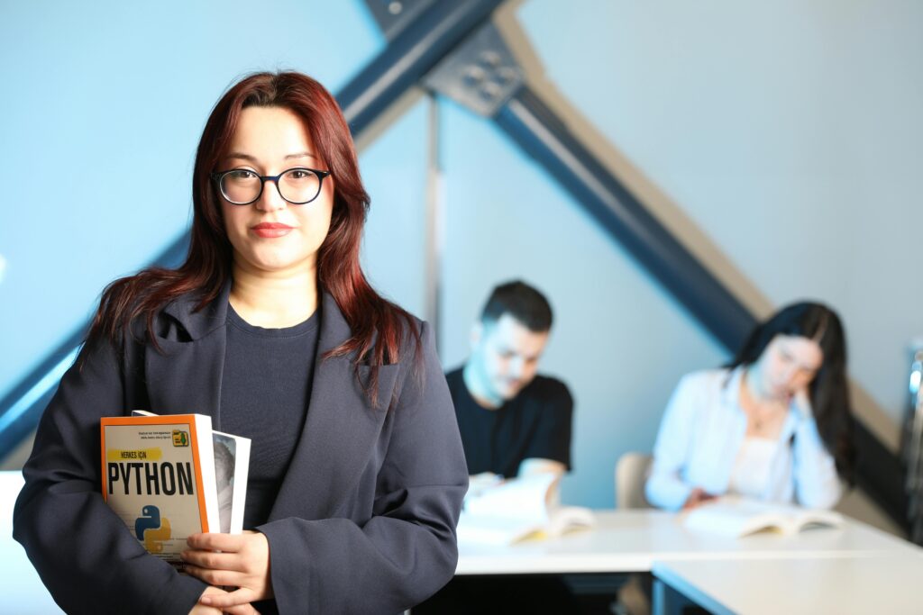 Woman in classroom setting holding Python programming book, with students in background.