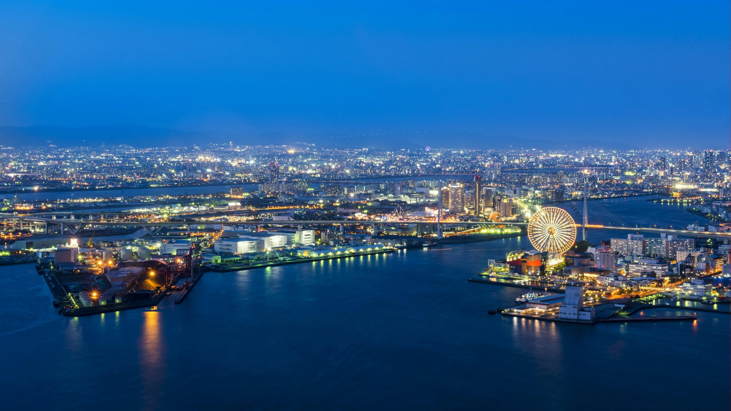 Aerial view of a vibrant city skyline by night featuring a glowing Ferris wheel and illuminated waterfront.