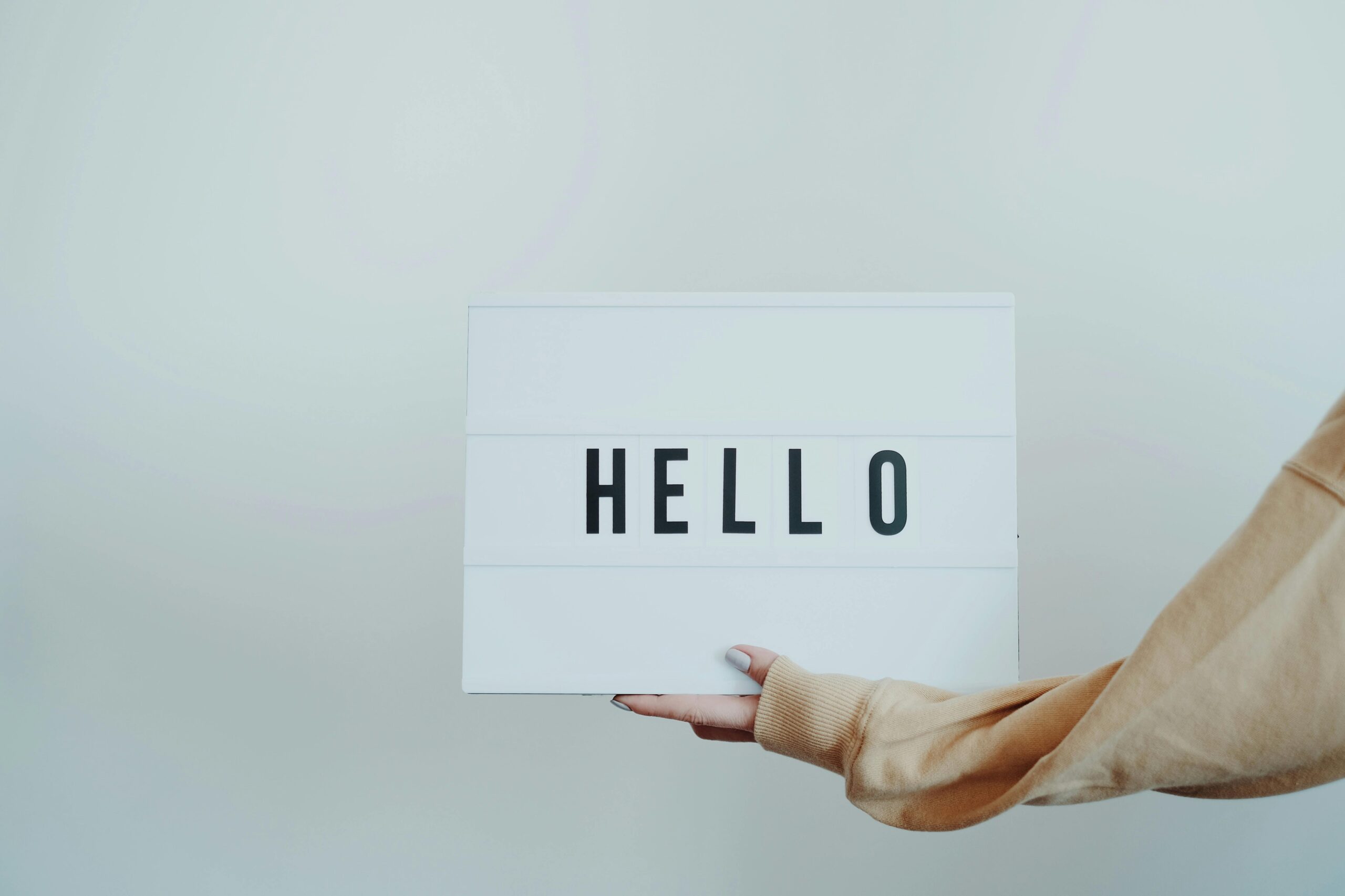 A hand holds a white sign displaying the word 'Hello' against a plain background.
