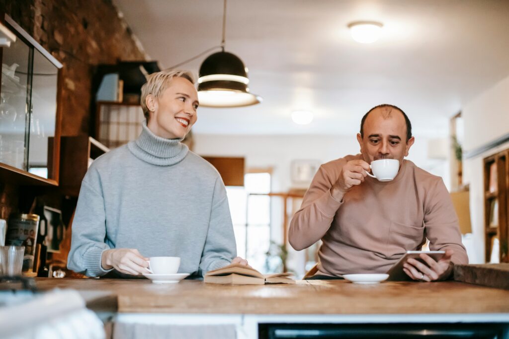 Happy adult couple at counter drinking tea from cups with saucer near opened book and while using tablet in light room