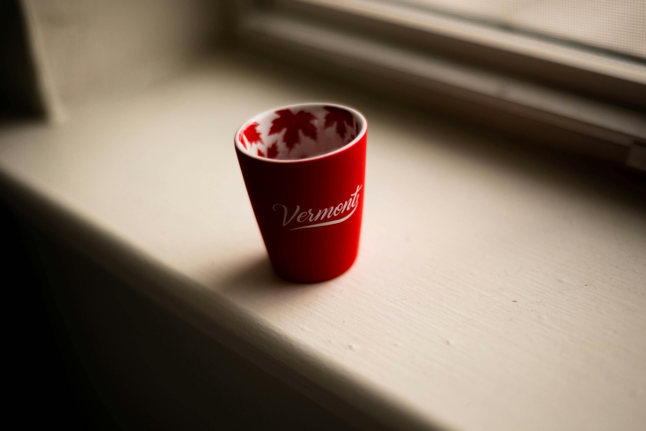 Close-up of a red Vermont mug with maple leaf design on a windowsill, soft natural light.