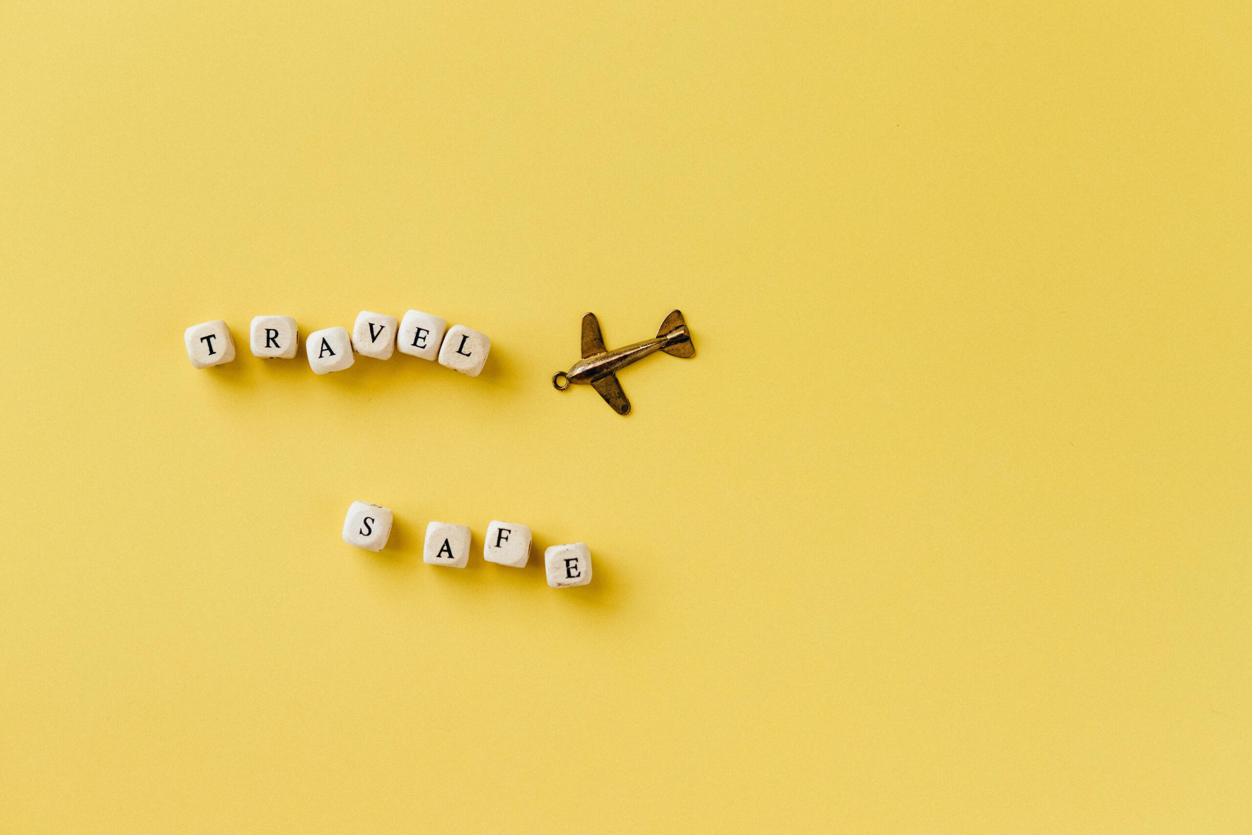 Flat lay of 'Travel Safe' text with airplane pendant on yellow background, evoking travel and safety concepts.