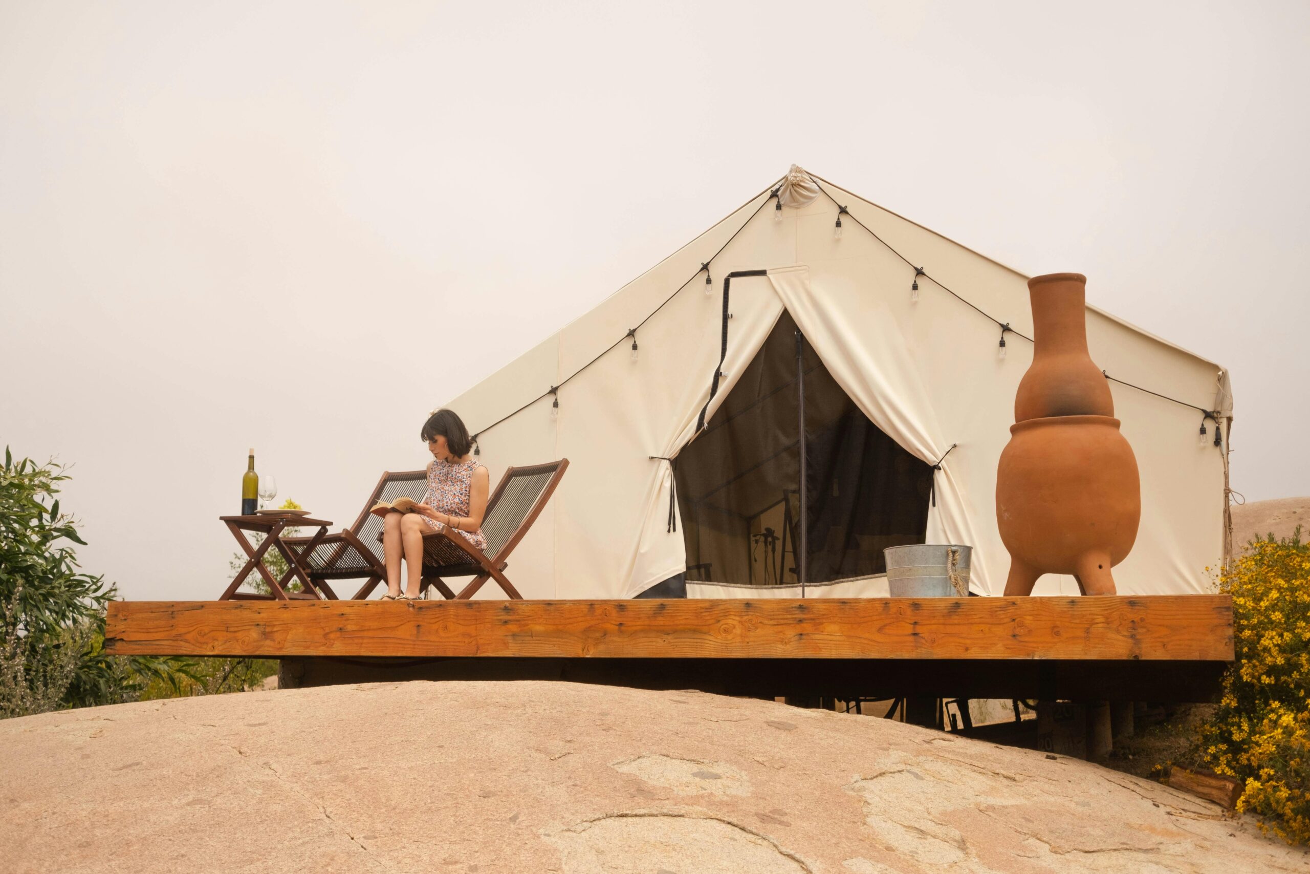 Woman reading outside a glamping tent with wine and nature ambiance.