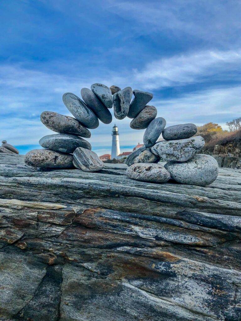 A serene image of balanced stones with the iconic Portland Head Lighthouse in the background.