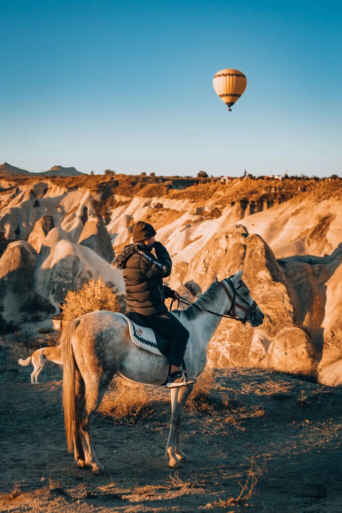 A horseback rider in Cappadocia observes a hot air balloon floating over unique rock formations.