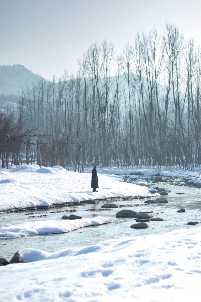 A lone figure stands by a snowy riverbank in Srinagar, surrounded by winter beauty.
