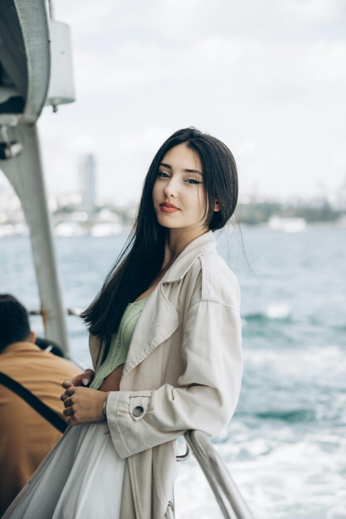 A young woman in a beige coat poses elegantly on a ferry with a scenic view of Istanbul in the background.