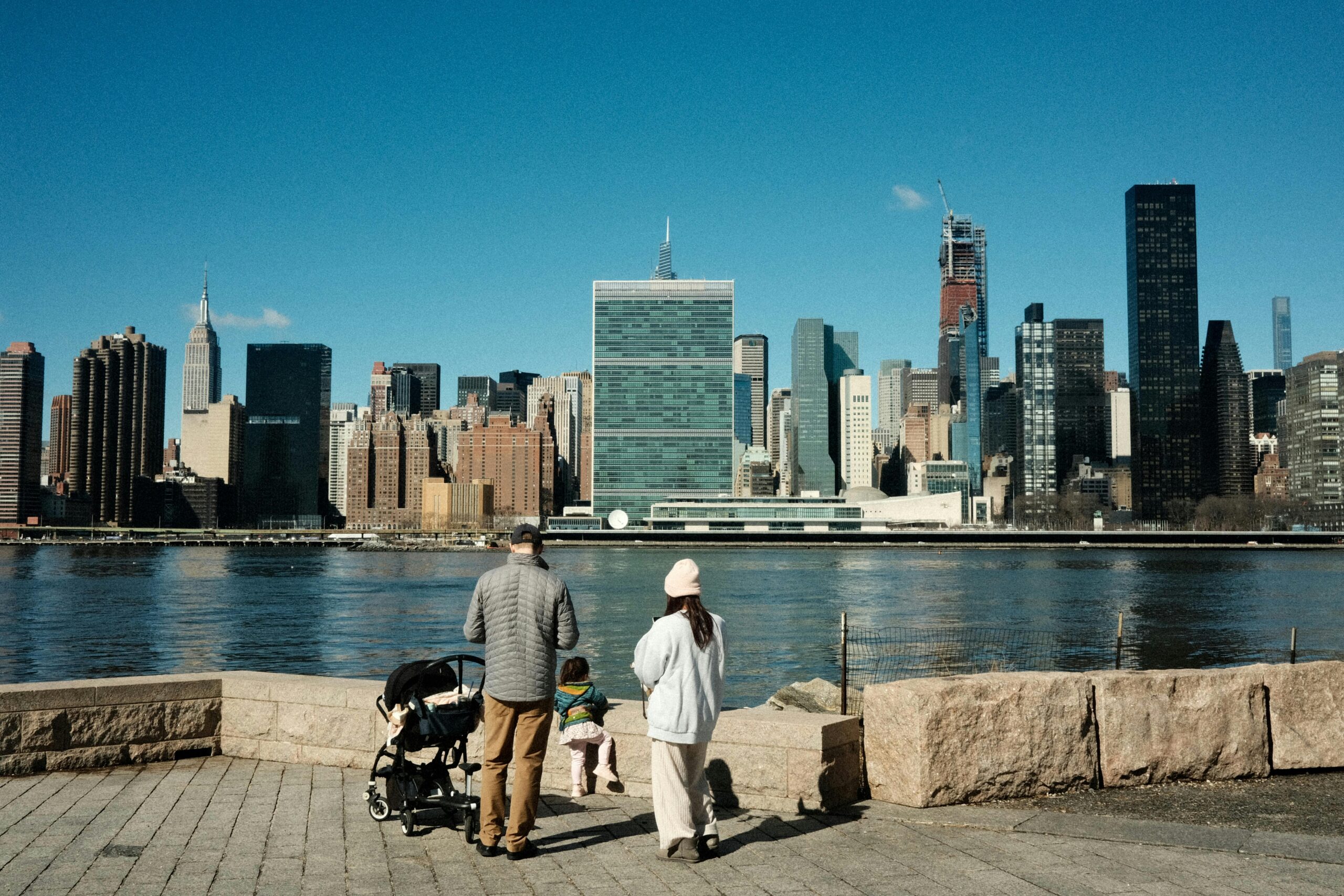 A family enjoys a riverside stroll with New York City's iconic skyline as the backdrop.