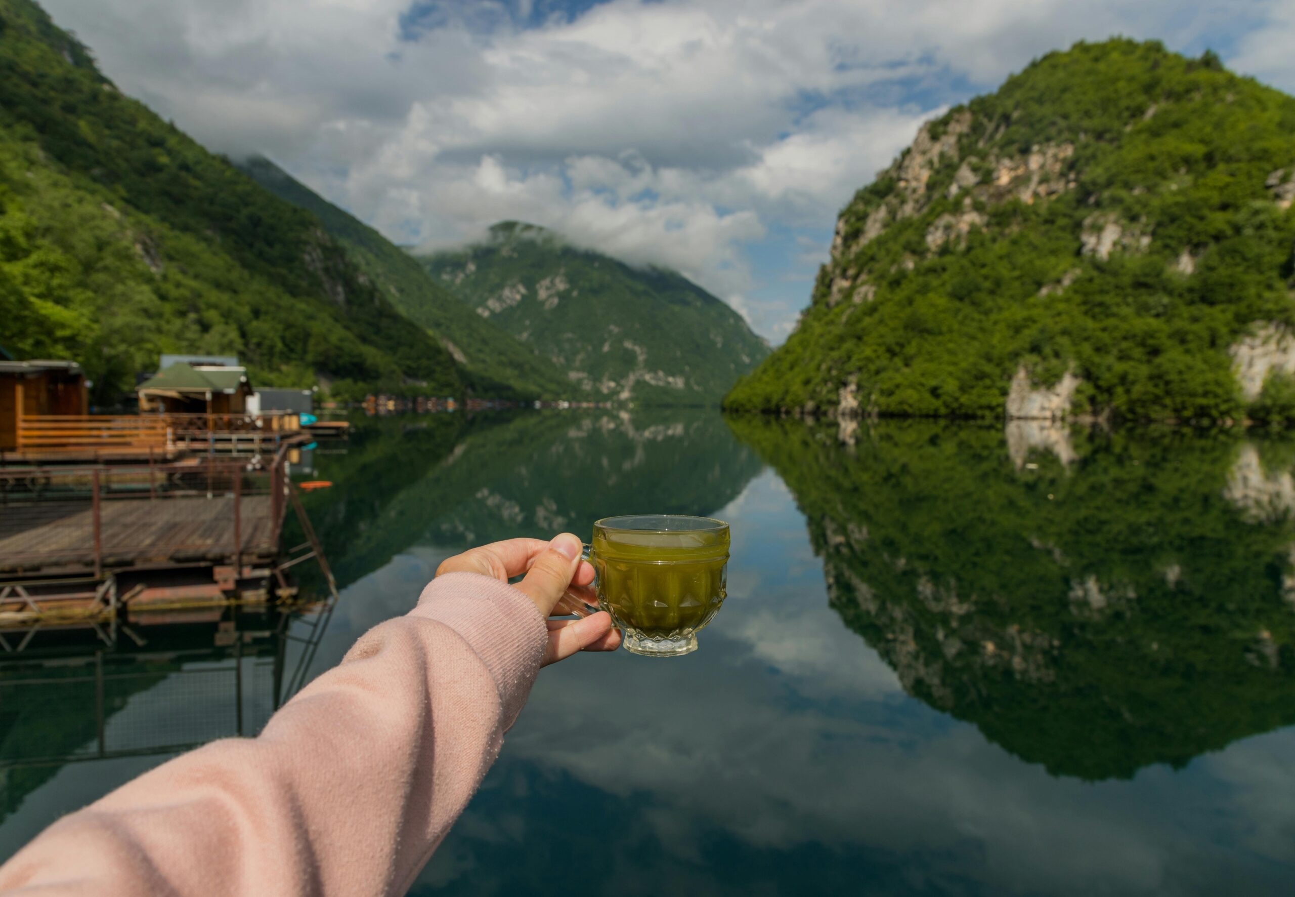 Scenic view of a woman holding a glass of matcha tea with a serene lake and lush hills in Perućac, Serbia.