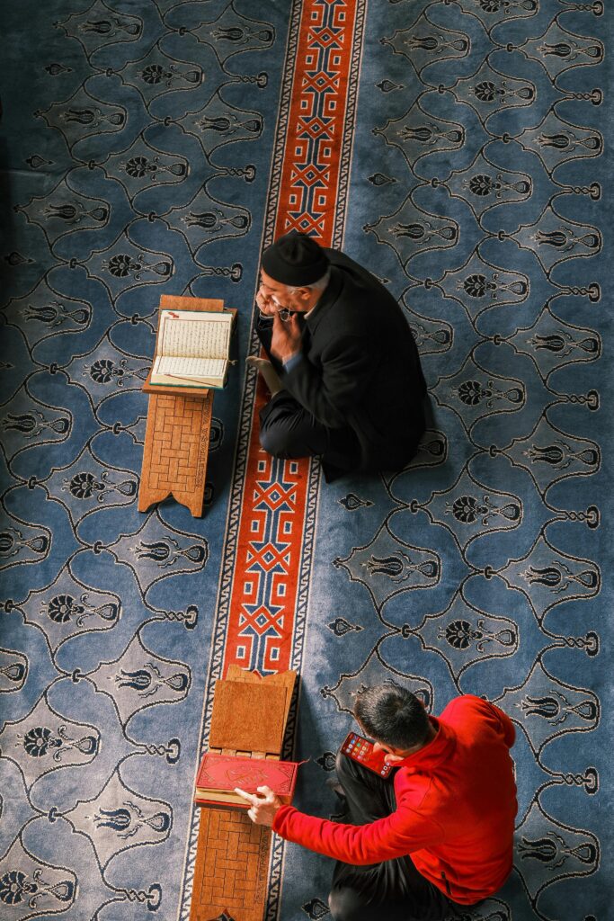 Two people in traditional attire reading in a mosque interior setting.