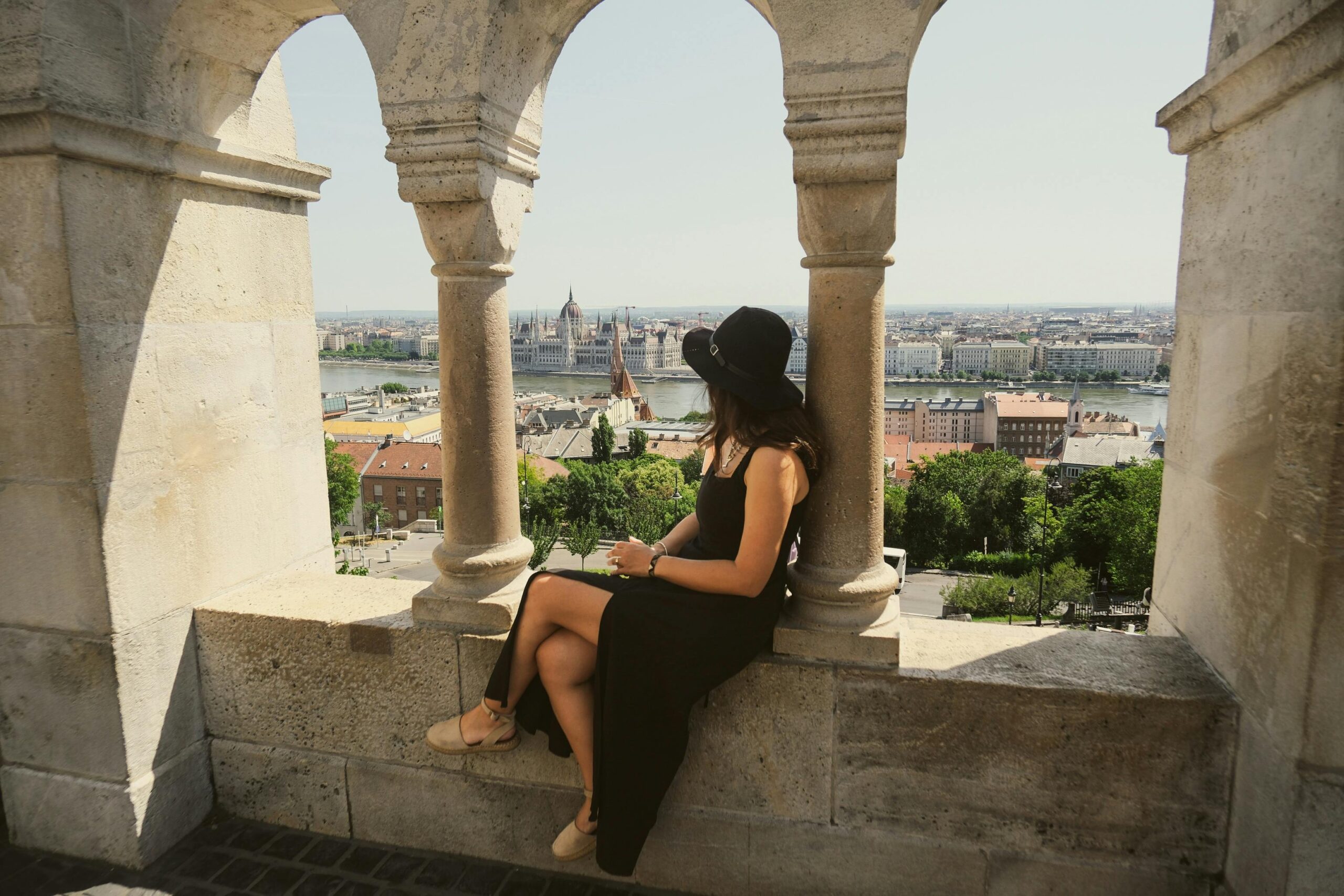 A woman in a black dress enjoys the panoramic view of Budapest from Fisherman's Bastion on a sunny day.