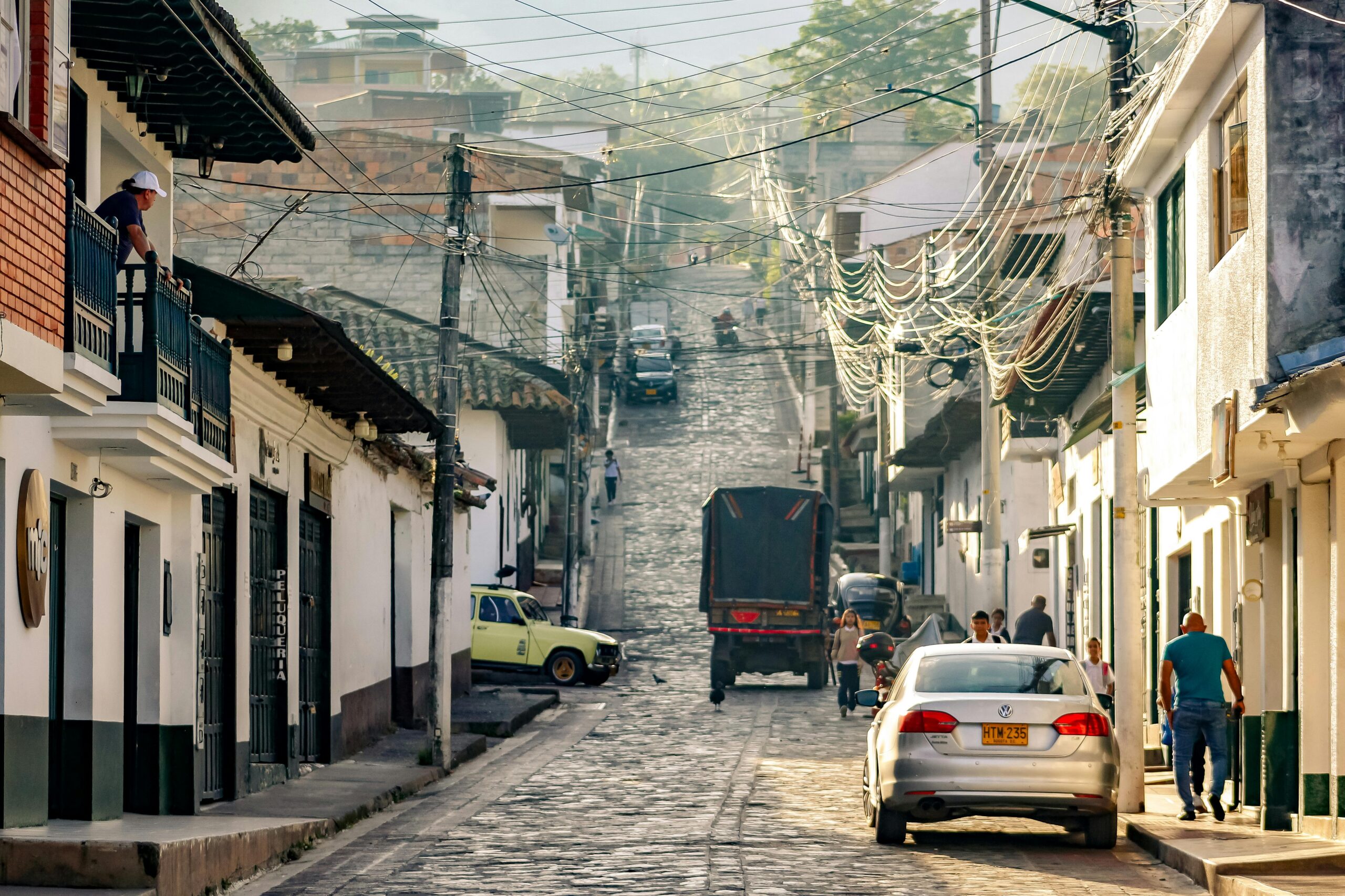 Street in a Colombian town with cobblestones, cars, and pedestrians, bathed in golden sunlight.