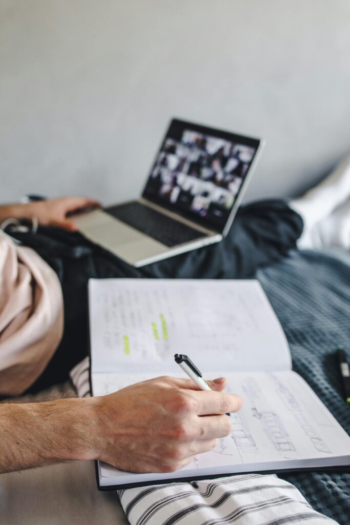 Student taking notes with a notebook during an online class on a laptop, a modern distance learning setup.