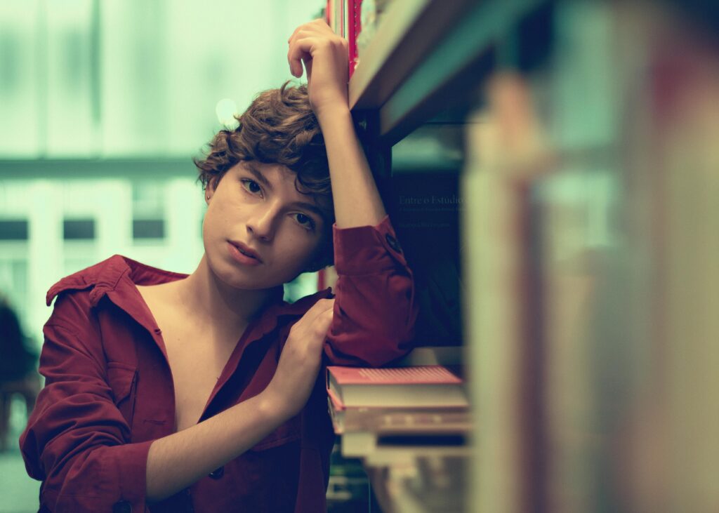 A thoughtful woman in a red shirt poses by bookshelves in a library.